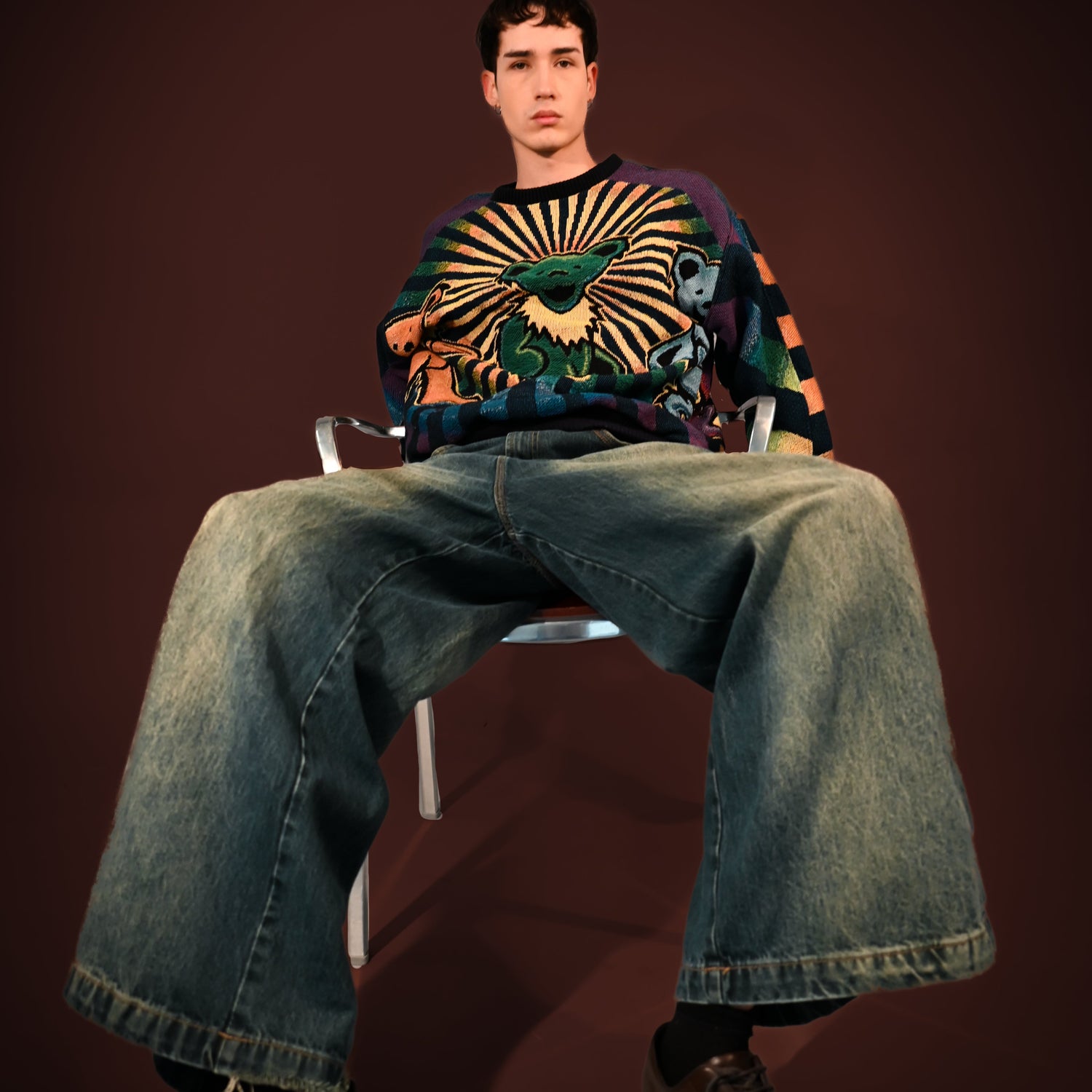 Person sitting on a chair wearing a colorful sweater and jeans against a dark background